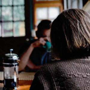 Kvinne med ryggen til kamera som sitter ved et kjøkkenbord overfor en mann som drikker kaffe.